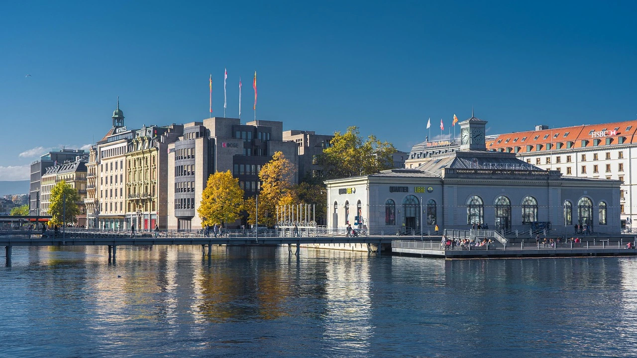 eine Stadtansicht, im Vordergrund ein See, im Hintergrund mehrere offizielle Gebäude mit Flaggen auf den Dächern
