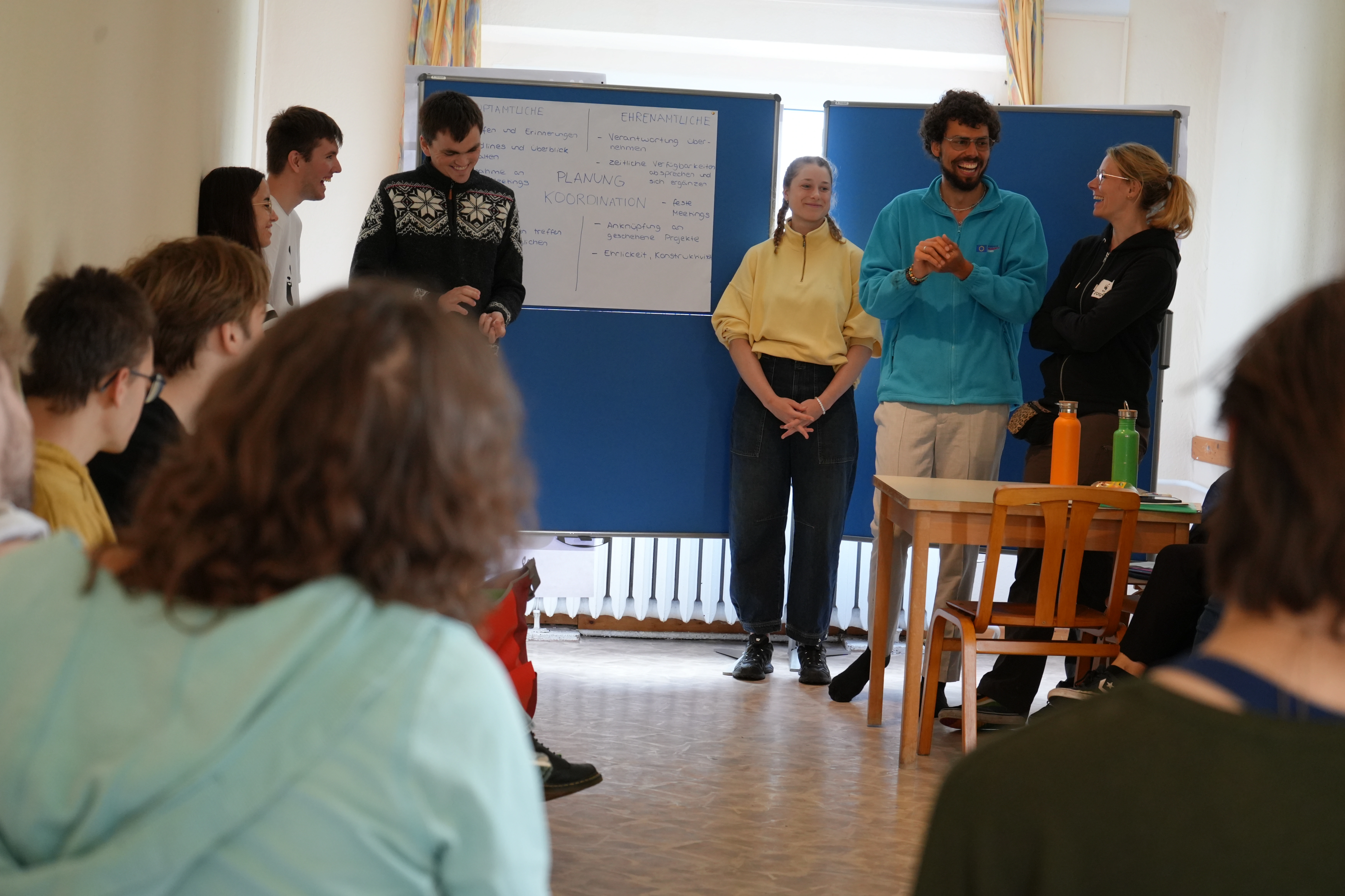 An einer Präsentationswand hängt ein Plakat "Planung und Koordination, Ehrenamtliche und Hauptamtliche" steht darauf mit Unterpunkten, die auf die Distanz nicht lesbar sind. Die Präsentierenden lachen.