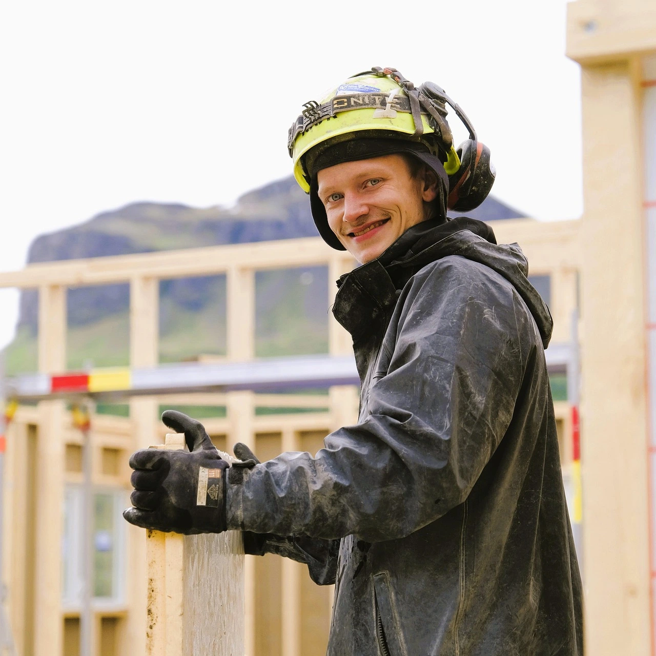 Young adult male on a construction site, with hard hard and ear defenders, holding a plank of wood