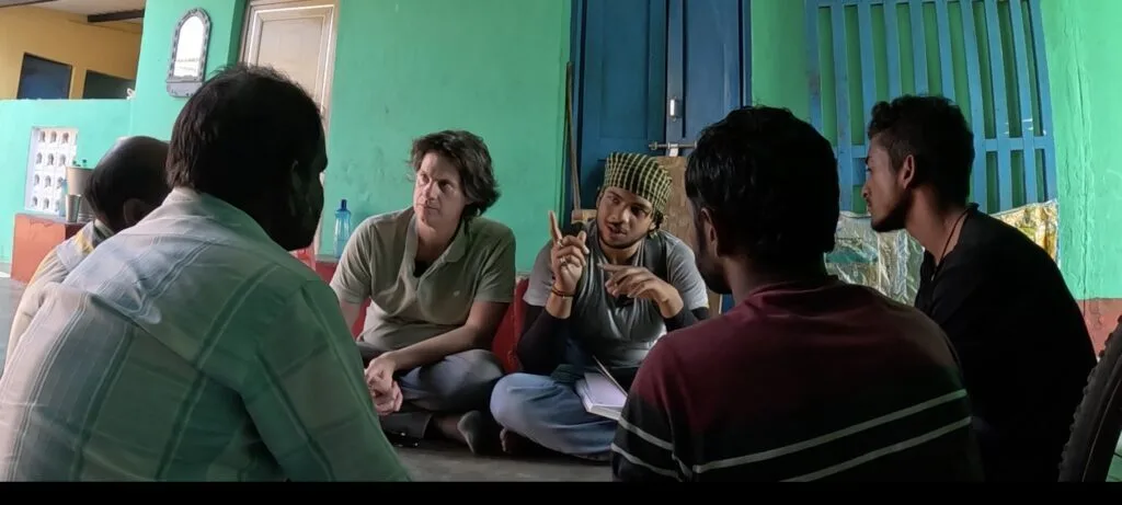 Jason Strother and a young man are sat cross-legged in a circle of four others, who have their backs to the camera. Jason is in discussion with the group for the Invisible Impact documentary in Odisha, eastern India.